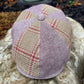 A eight-panel flat cap hat made from textured tweed fabric, featuring a mix of dusty lavender, beige, and red plaid panels, resting on a large stone outdoors.