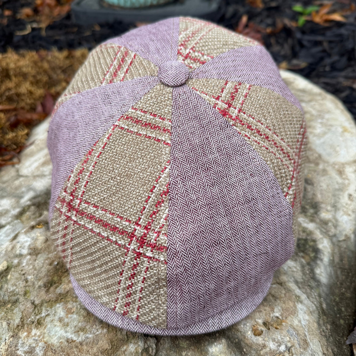 A eight-panel flat cap hat made from textured tweed fabric, featuring a mix of dusty lavender, beige, and red plaid panels, resting on a large stone outdoors.