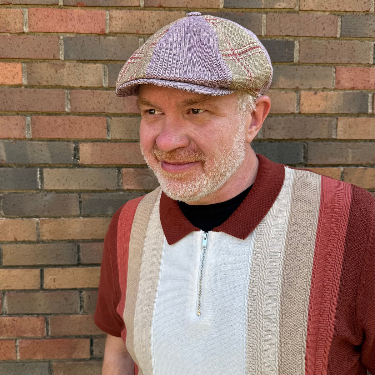 A smiling older man wearing a patchwork flat cap and a striped knit polo shirt, standing in front of a brick wall.