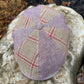 A structured flat cap made from lavender and beige woven fabric with red plaid lines, resting on a large stone outdoors.