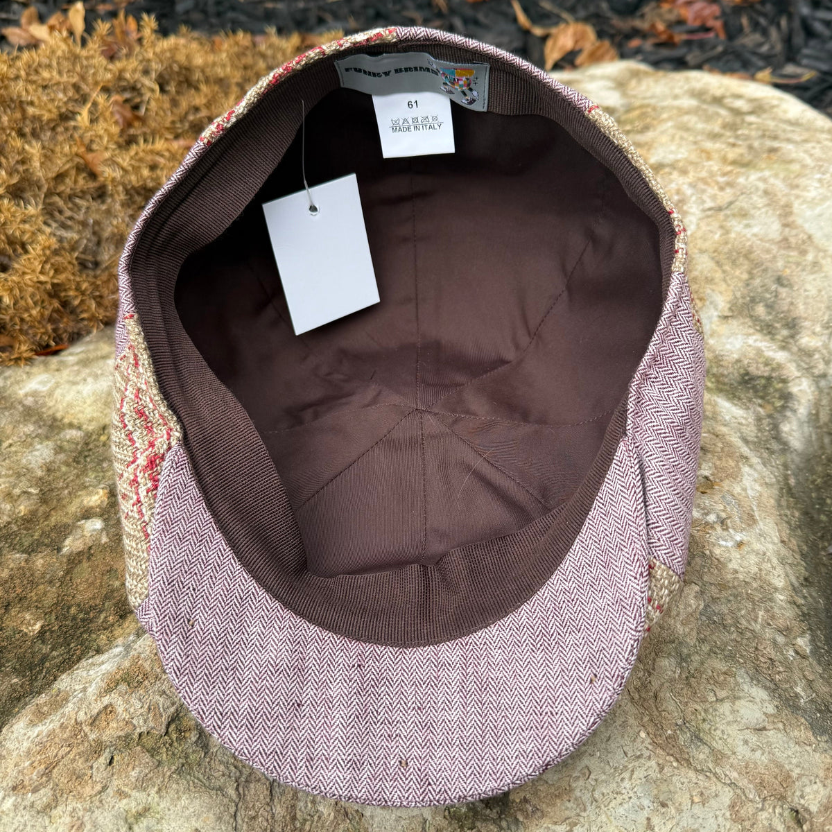 A flat-brim cap with a dusty rose and brown herringbone patterned exterior and a dark brown interior, resting on a large stone outdoors.