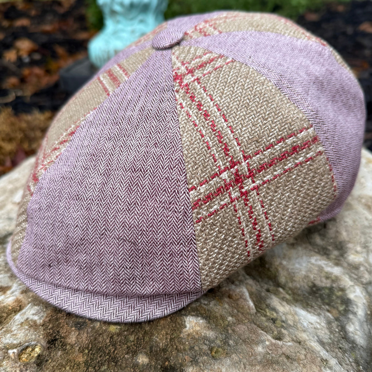 A eight-panel flat cap hat made from a patchwork of tweed fabrics, featuring a dusty lavender herringbone pattern, a beige and red plaid panel, and a matching lavender chevron panel, resting on a textured stone.