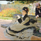 Impressionist oil painting depicting a woman seated on a bench in a park, with a man leaning on the bench behind her and flowers in the foreground.