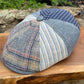 A patchwork flat cap made from multiple woven fabrics, including plaid, striped, and houndstooth patterns in blue, gray, beige, and red, resting on a wooden surface with a blurred outdoor garden background.