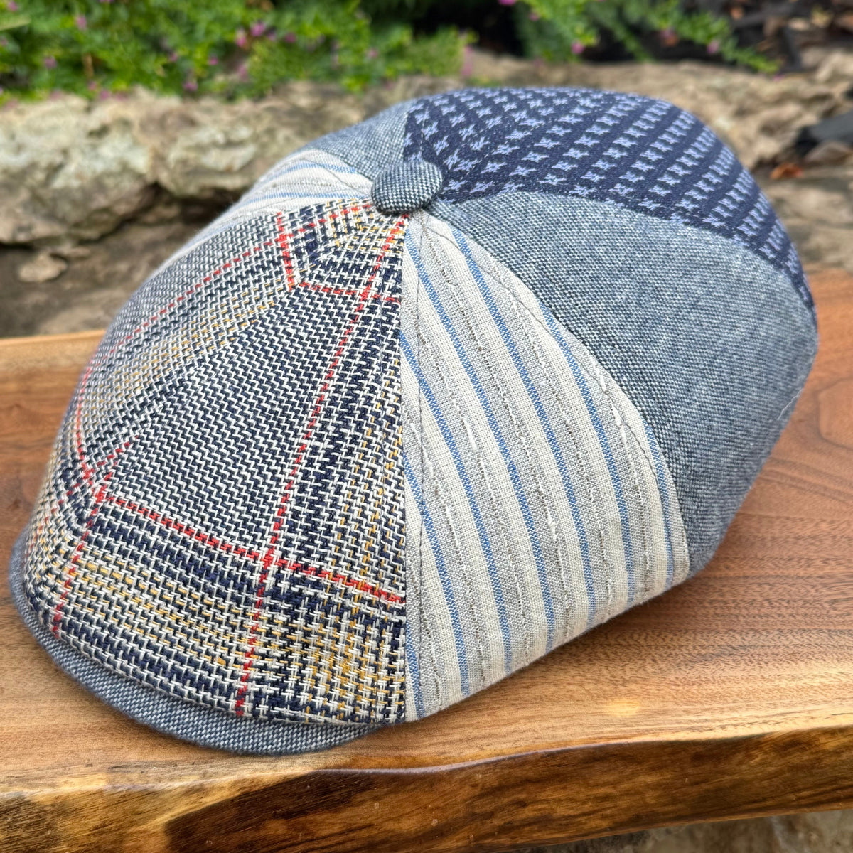 A patchwork flat cap made from multiple woven fabrics, including plaid, striped, and houndstooth patterns in blue, gray, beige, and red, resting on a wooden surface with a blurred outdoor garden background.