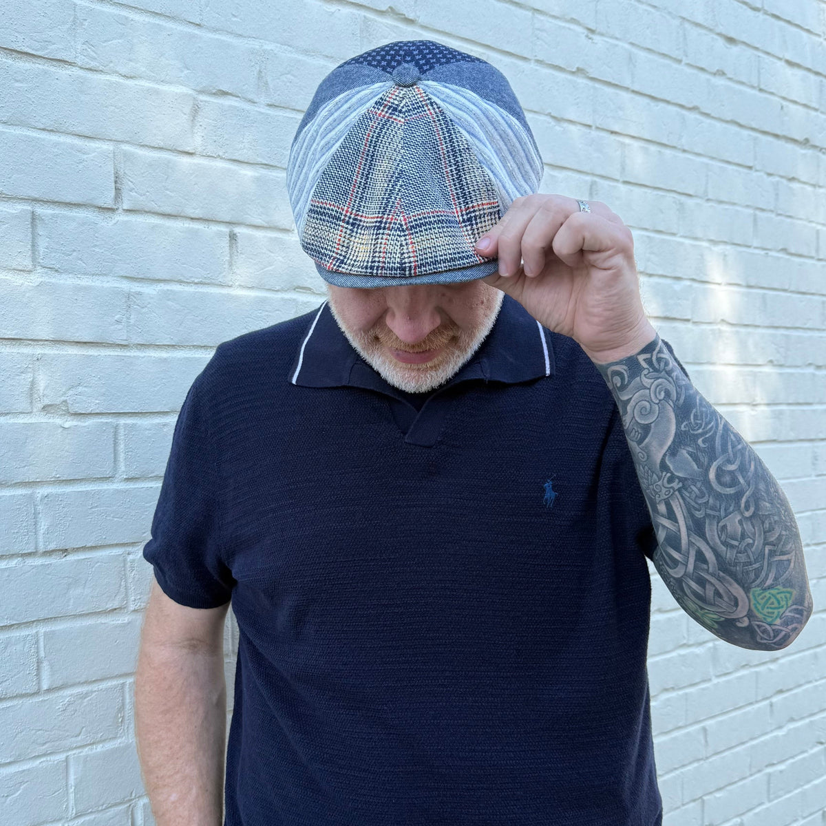 A man wearing a navy blue polo shirt and a blue and white plaid flat cap, standing against a white painted brick wall.