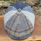 A patchwork flat cap made from various textured fabrics, including a navy blue star-patterned section, a light blue striped section, and a plaid tweed front panel with red, yellow, and blue lines. The cap is displayed on a wooden board with a blurred outdoor stone background.