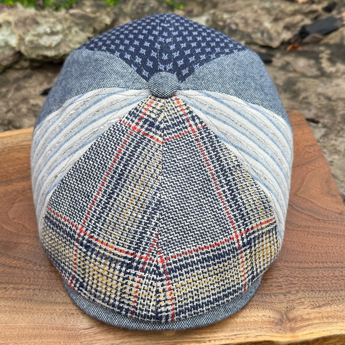A patchwork flat cap made from various textured fabrics, including a navy blue star-patterned section, a light blue striped section, and a plaid tweed front panel with red, yellow, and blue lines. The cap is displayed on a wooden board with a blurred outdoor stone background.