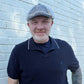 A man wearing a navy blue polo shirt with white piping, beige pants, and a blue plaid flat cap, standing against a white painted brick wall.
