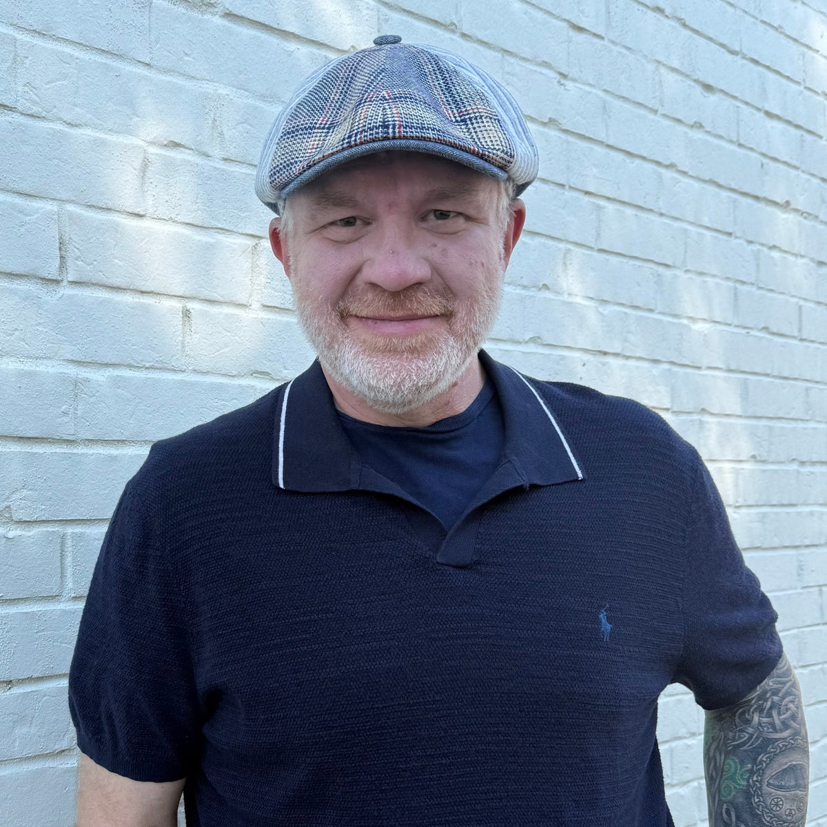 A man wearing a navy blue polo shirt with white piping, beige pants, and a blue plaid flat cap, standing against a white painted brick wall.