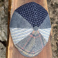 A handmade patchwork flat cap made from multiple fabric panels, displayed on a rustic wooden board with a stone background. The cap features a navy blue houndstooth pattern on the top panel, solid blue denim panels on the sides, light blue and white striped fabric on the front-left, and a plaid pattern with blue, beige, and red lines on the front-right.
