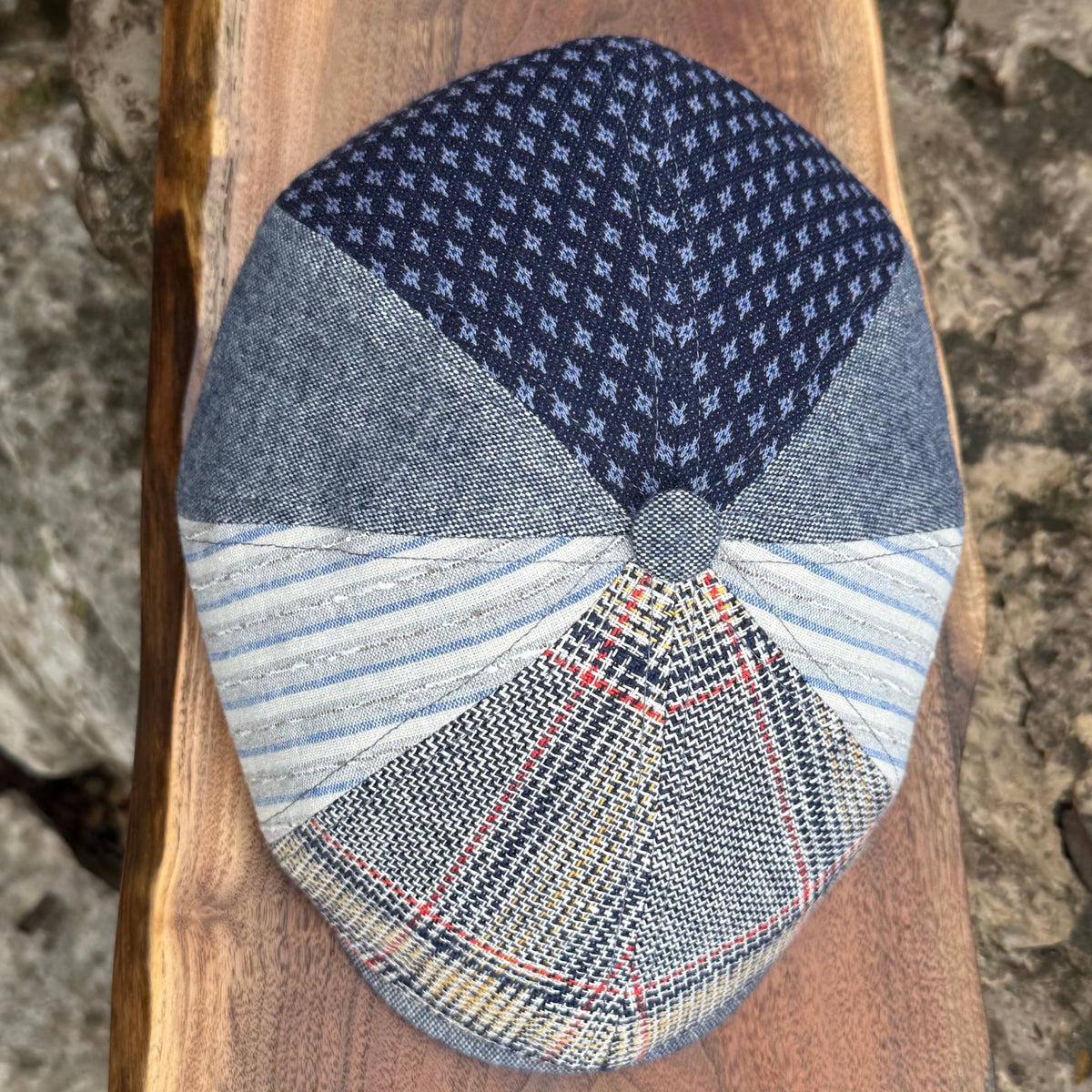A handmade patchwork flat cap made from multiple fabric panels, displayed on a rustic wooden board with a stone background. The cap features a navy blue houndstooth pattern on the top panel, solid blue denim panels on the sides, light blue and white striped fabric on the front-left, and a plaid pattern with blue, beige, and red lines on the front-right.