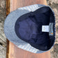 A close-up top-down view of a denim flat cap resting on a wooden surface, showing the blue-gray denim fabric, striped side panels, dark navy interior, and a sewn-in size label.