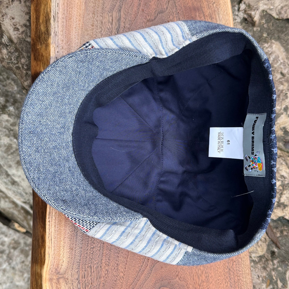 A close-up top-down view of a denim flat cap resting on a wooden surface, showing the blue-gray denim fabric, striped side panels, dark navy interior, and a sewn-in size label.