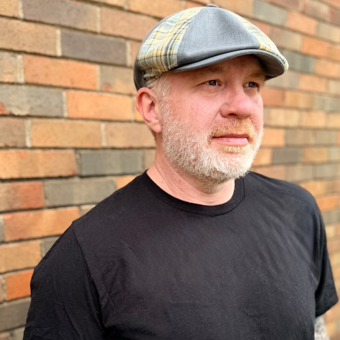 Man wearing a black t-shirt and flat cap standing against a brick wall.