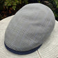 A gray and black houndstooth flat cap with a navy blue textured brim, resting on a light-colored stone surface with green garden foliage in the background.