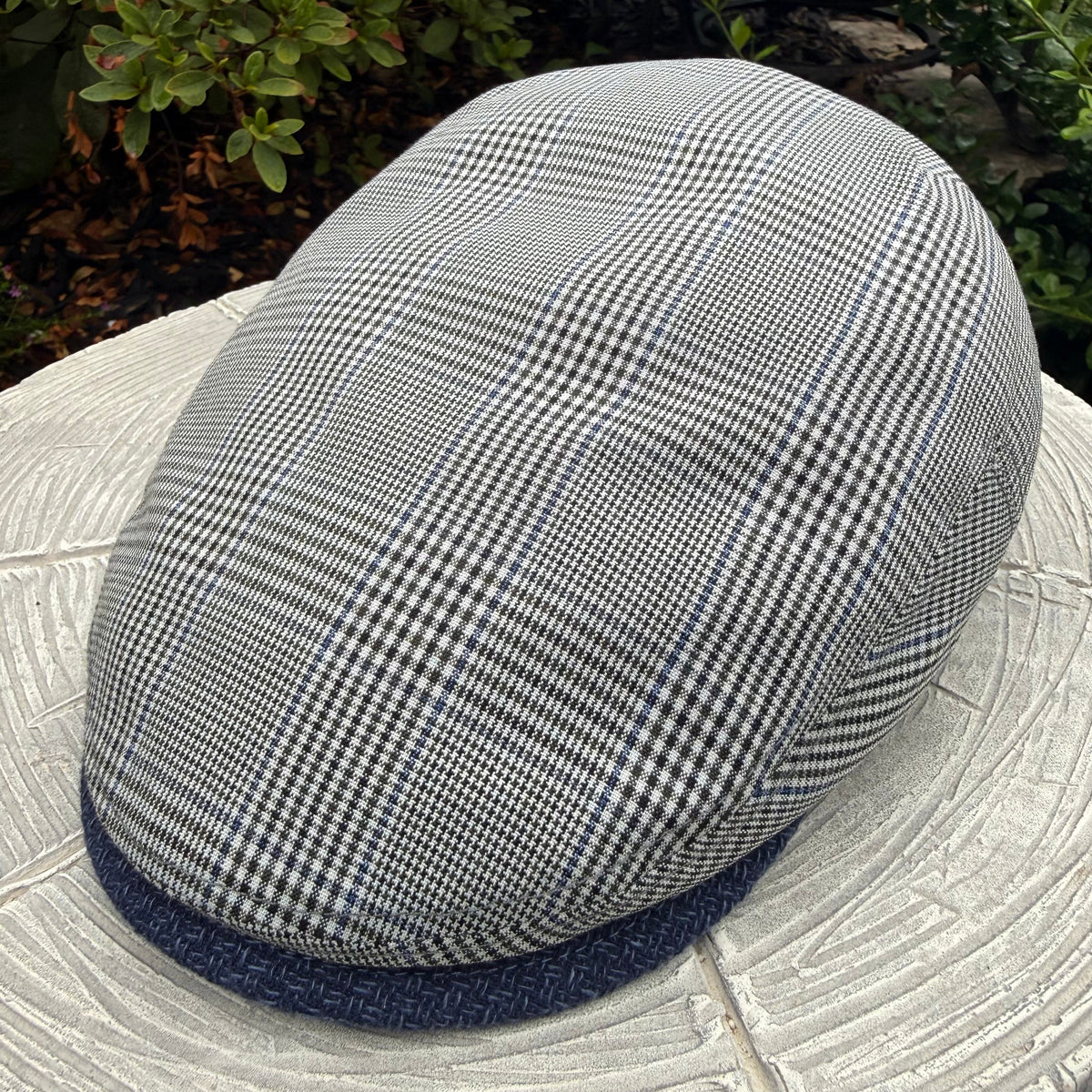 A gray and black houndstooth flat cap with a navy blue textured brim, resting on a light-colored stone surface with green garden foliage in the background.