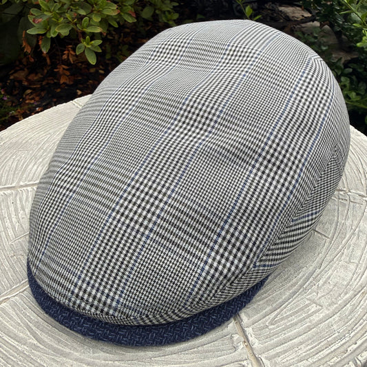 A gray and black houndstooth flat cap with a navy blue textured brim, resting on a light-colored stone surface with green garden foliage in the background.