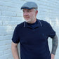A man wearing a navy blue polo shirt with white piping on the collar and a gray patterned flat cap, standing against a white painted brick wall.
