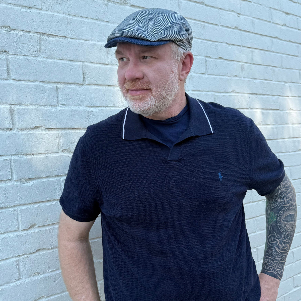 A man wearing a navy blue polo shirt with white piping on the collar and a gray patterned flat cap, standing against a white painted brick wall.