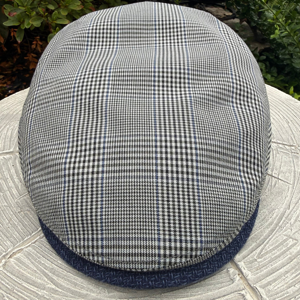 A classic flat cap with a black and white houndstooth pattern and a navy blue textured brim, photographed outdoors on a stone surface with green foliage in the background.