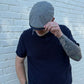 A man wearing a navy blue short-sleeve polo shirt and a gray plaid flat cap, standing against a white painted brick wall.