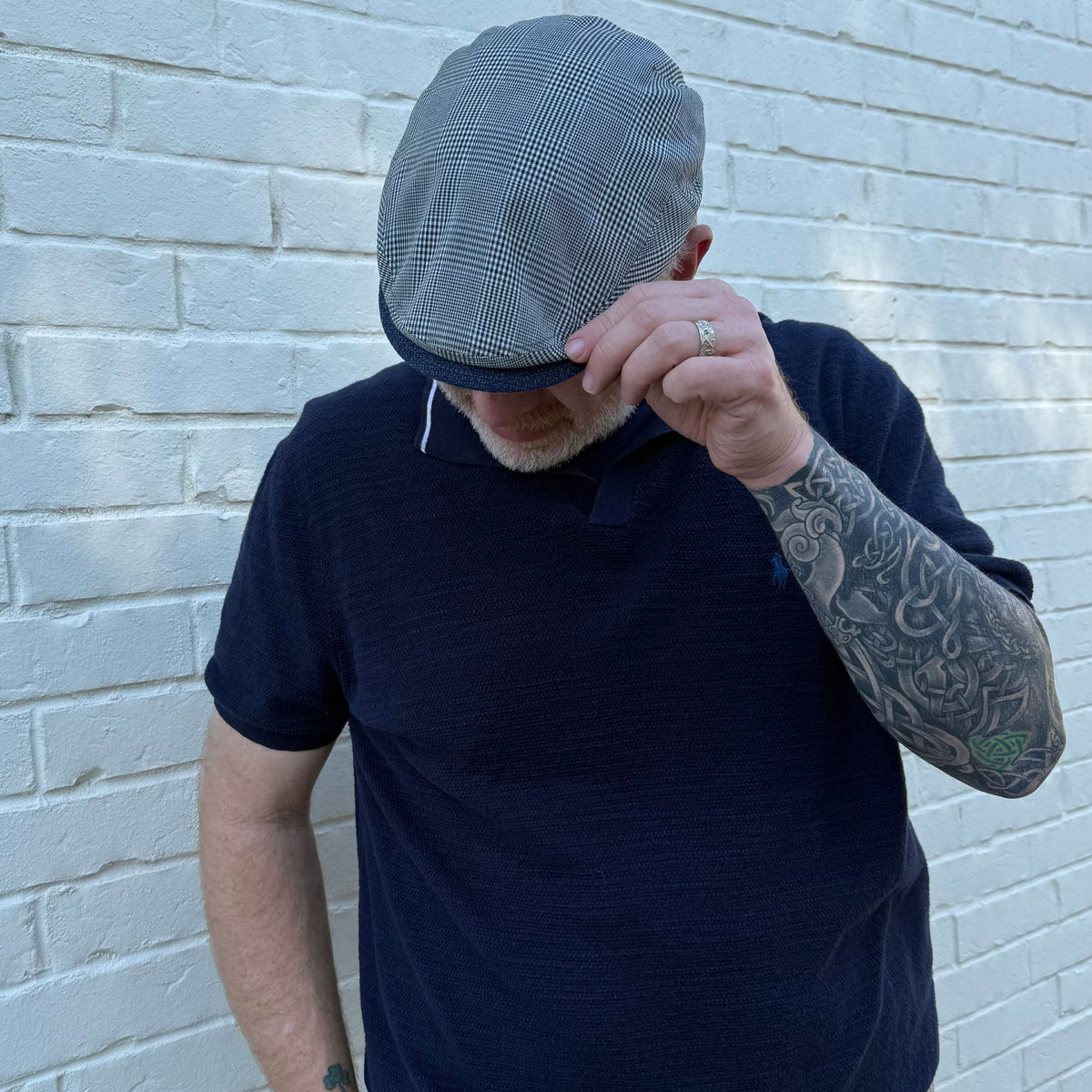 A man wearing a navy blue short-sleeve polo shirt and a gray plaid flat cap, standing against a white painted brick wall.