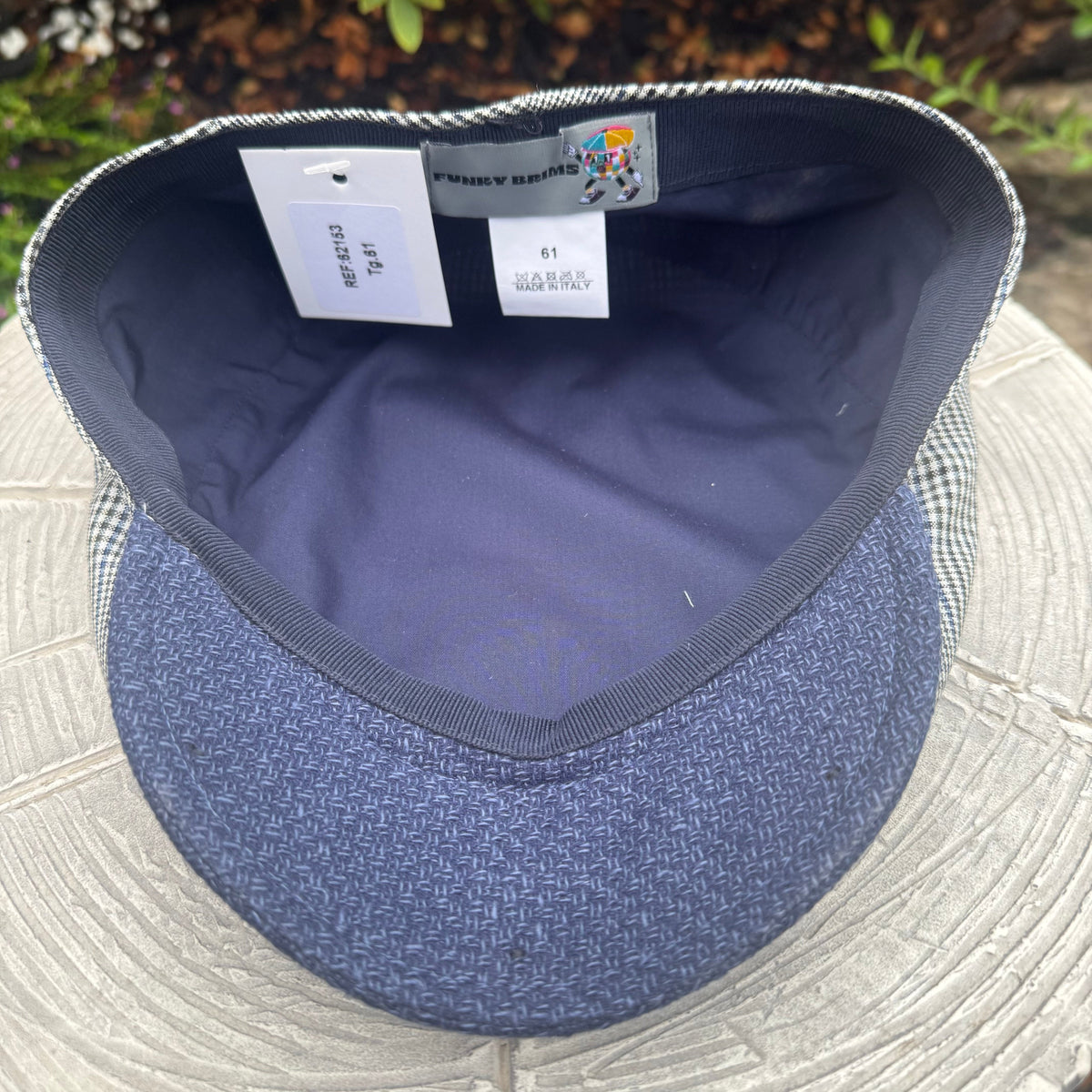 A blue herringbone patterned flat cap with a navy blue interior, resting on a light gray stone surface. The brand label 'Funky Hats' is visible inside the crown.