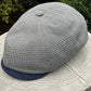 A structured flat cap with a small-scale black and white gingham pattern on the crown and a solid navy blue visor, resting on a light-colored stone surface with green foliage in the background.