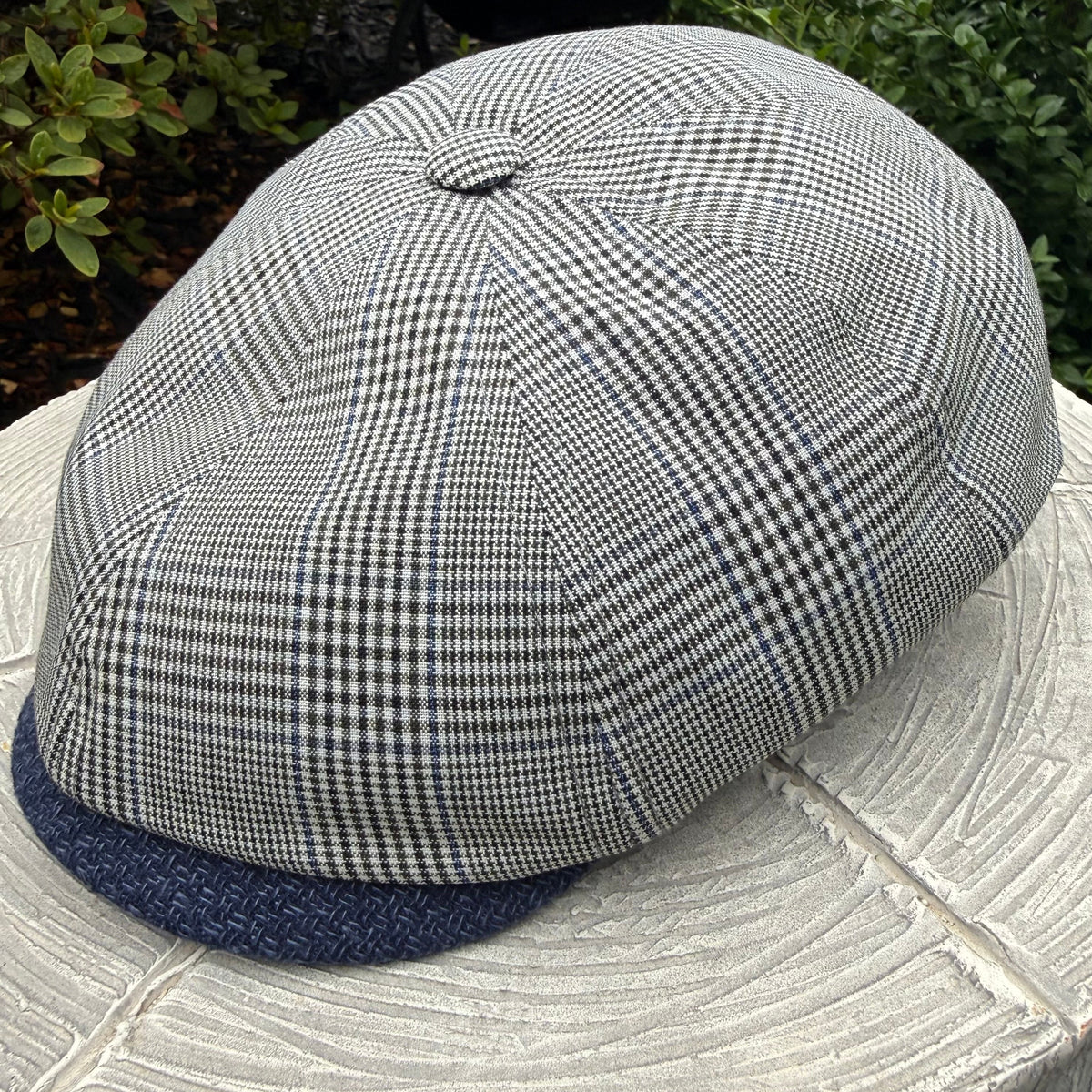A structured flat cap with a small-scale black and white gingham pattern on the crown and a solid navy blue visor, resting on a light-colored stone surface with green foliage in the background.