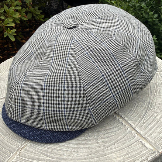 A structured flat cap with a small-scale black and white gingham pattern on the crown and a solid navy blue visor, resting on a light-colored stone surface with green foliage in the background.