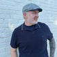 A man wearing a navy blue polo shirt with white piping and a gray flat cap, standing against a white painted brick wall.