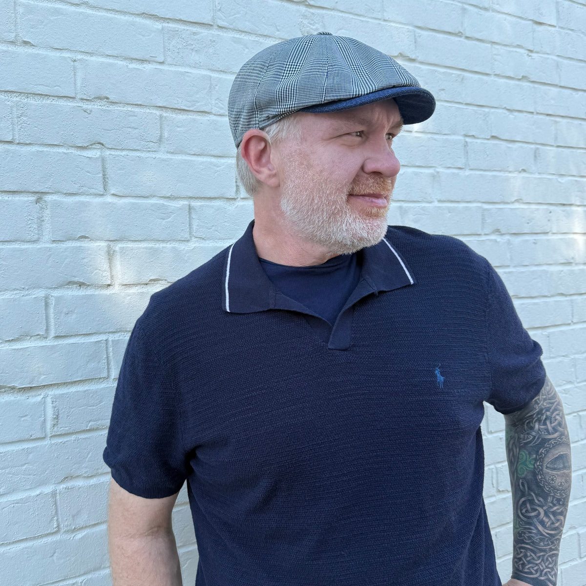 A man wearing a navy blue polo shirt with white piping and a gray flat cap, standing against a white painted brick wall.