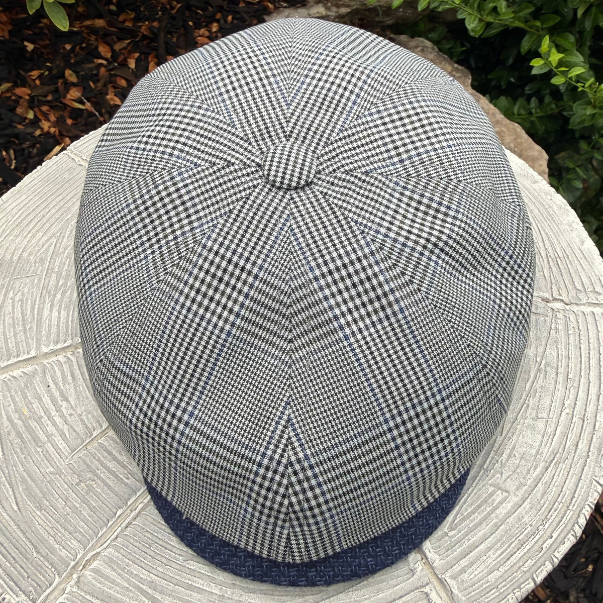 A top-down view of a classic flat cap with a black and white gingham pattern and a navy blue visor, resting on a textured white stone surface outdoors.