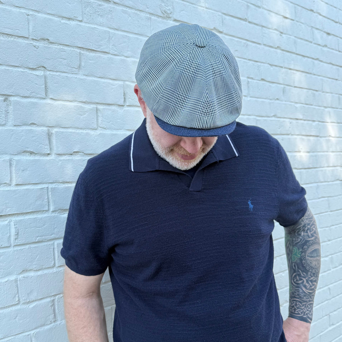 A man wearing a navy blue polo shirt with white piping and a gray checkered flat cap, standing against a white painted brick wall.