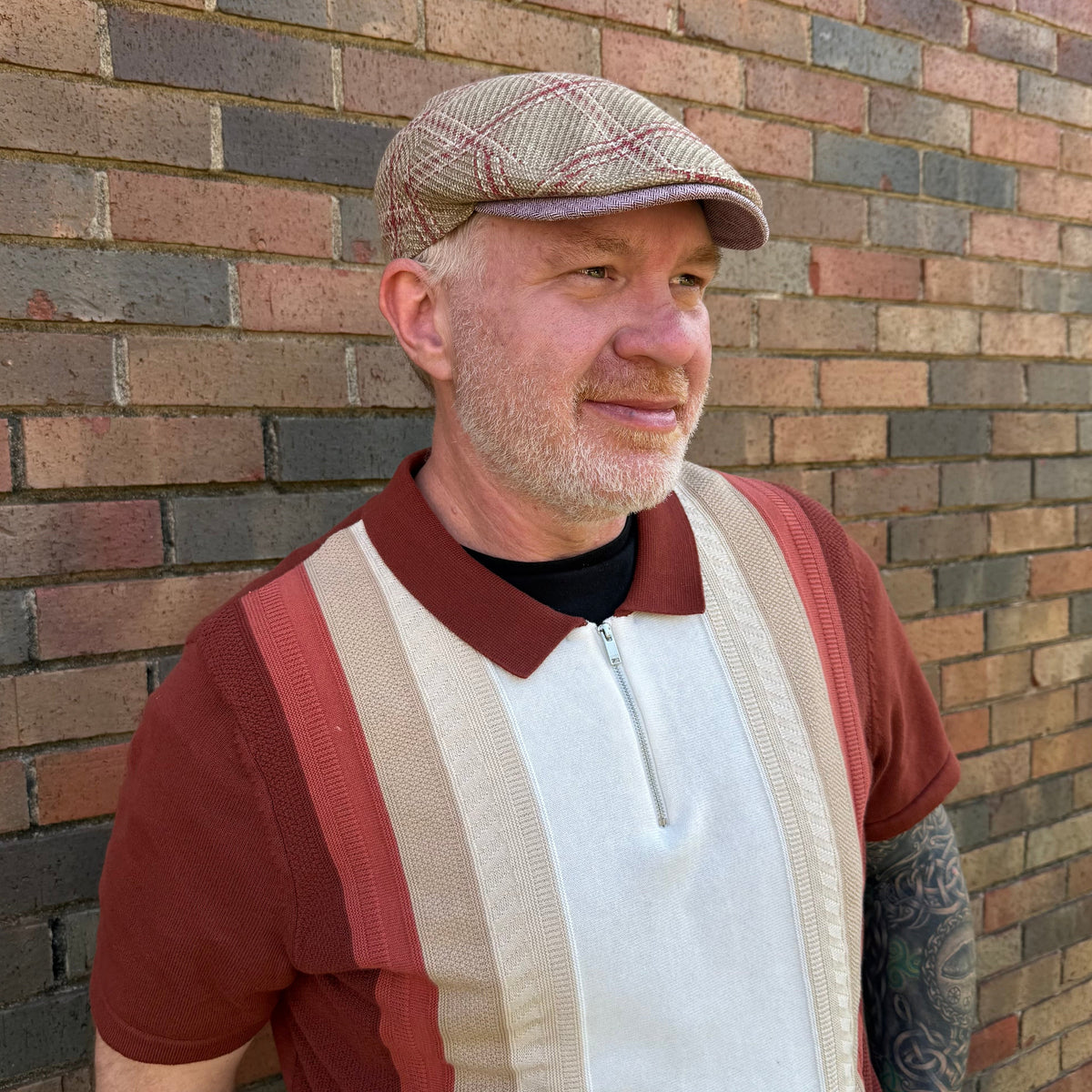 A man wearing a brown and beige herringbone flat cap, a rust red and beige striped knit polo shirt over a black t-shirt, standing against a brick wall.