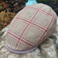 A woven flat cap in beige tweed with a red and white windowpane plaid pattern, resting on a large stone outdoors.