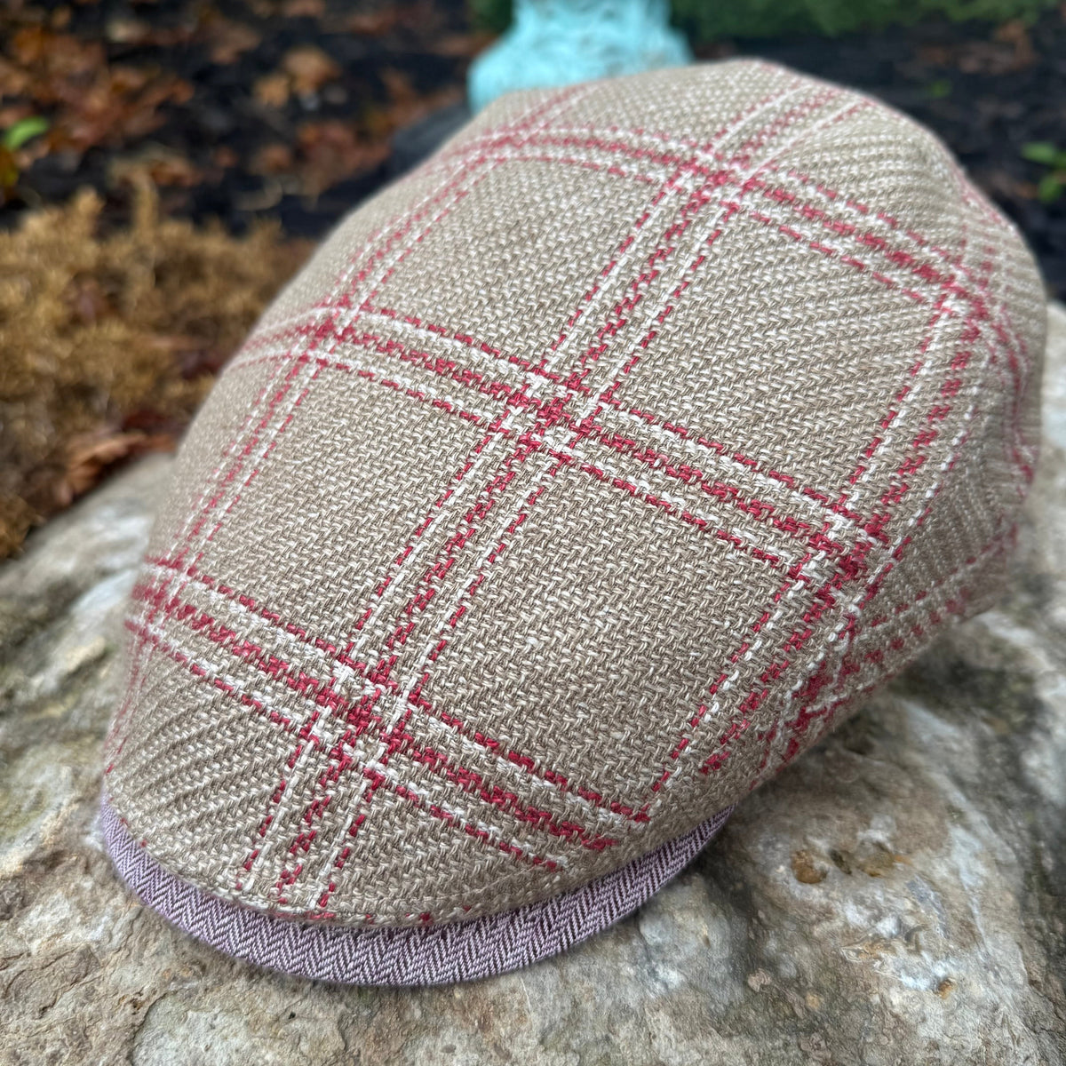 A woven flat cap in beige tweed with a red and white windowpane plaid pattern, resting on a large stone outdoors.