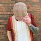 A man wearing a beige tweed flat cap with a red plaid pattern, a rust-colored knit sweater vest, and a cream knit sweater, standing in front of a brick wall.