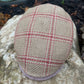 A close-up of a tweed flat cap resting on a large stone outdoors. The cap features a beige and brown woven base with a red windowpane plaid pattern and a matching red chevron trim along the edge.