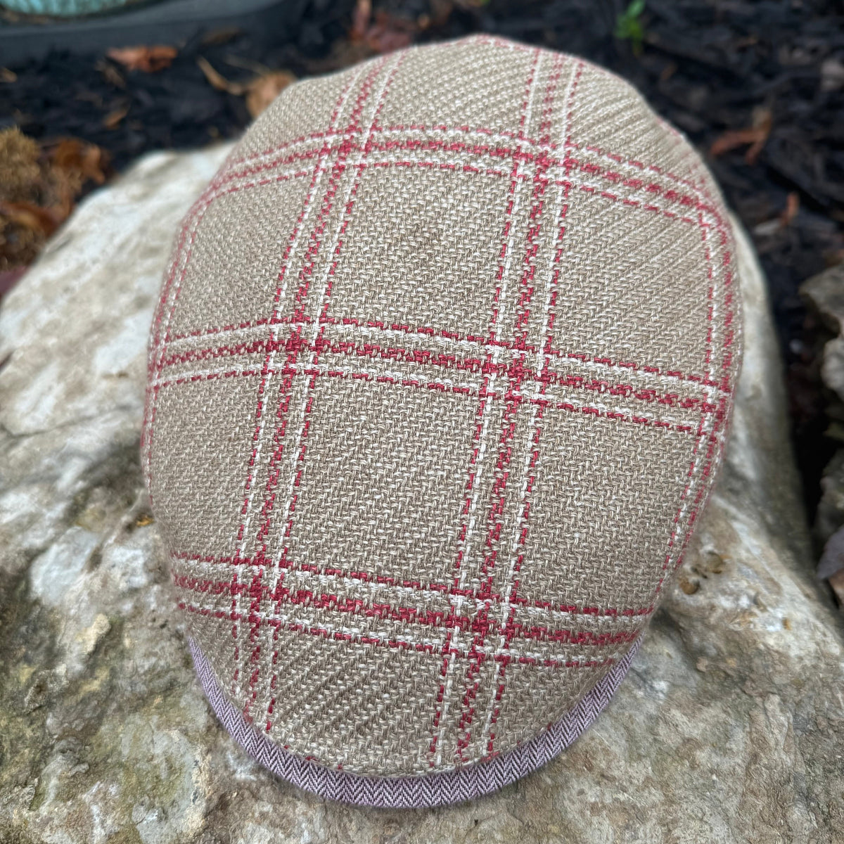 A close-up of a tweed flat cap resting on a large stone outdoors. The cap features a beige and brown woven base with a red windowpane plaid pattern and a matching red chevron trim along the edge.