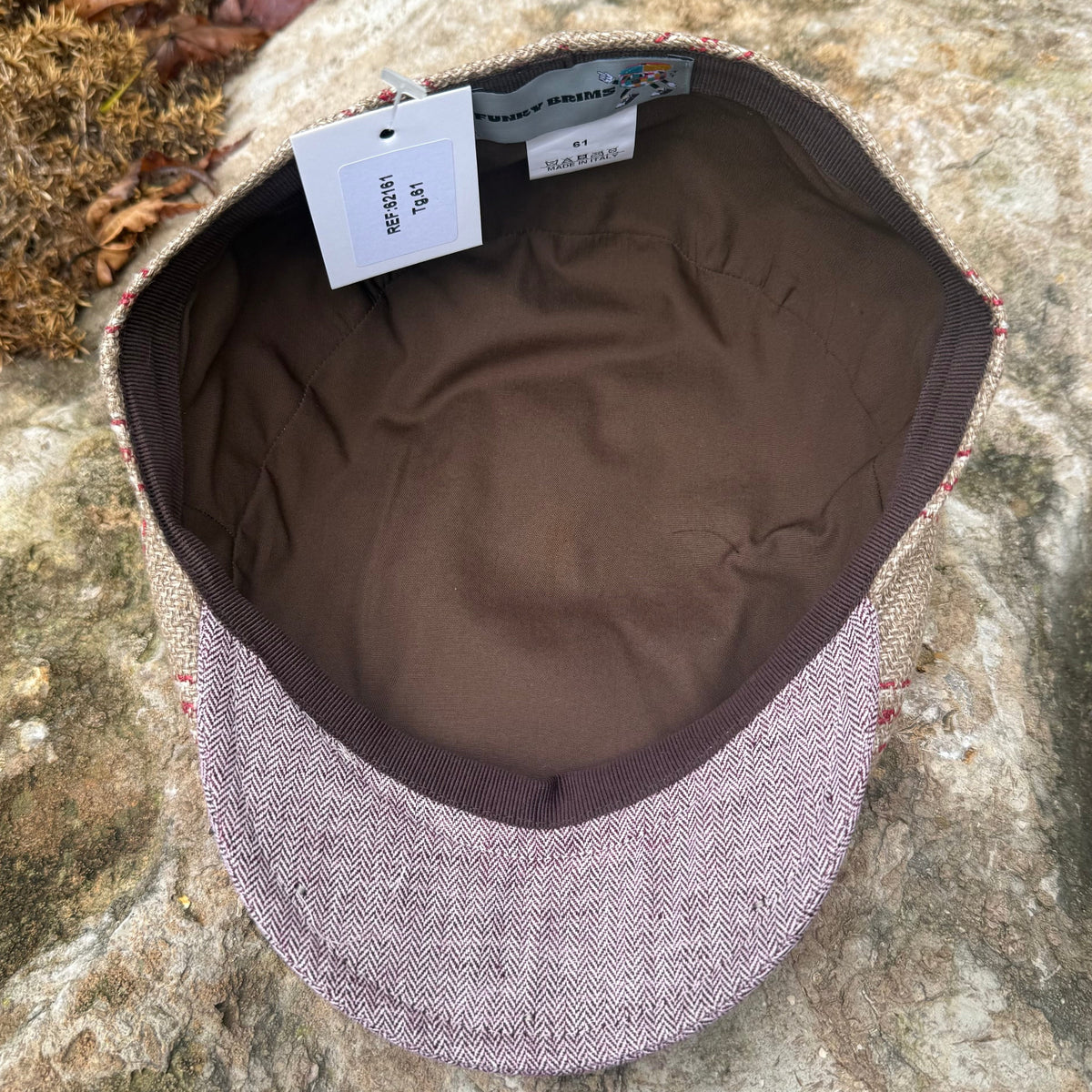 A flat-brim cap with a textured tweed exterior in muted pinkish-beige and brown, featuring a solid brown fabric interior and a white size tag, photographed outdoors on a large rock.
