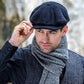 A man wearing a black newsboy cap, a gray and black herringbone scarf, and a matching tweed waistcoat over a black shirt, posed against a blurred natural stone background.