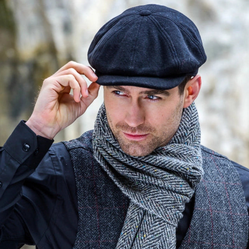 A man wearing a black newsboy cap, a gray and black herringbone scarf, and a matching tweed waistcoat over a black shirt, posed against a blurred natural stone background.