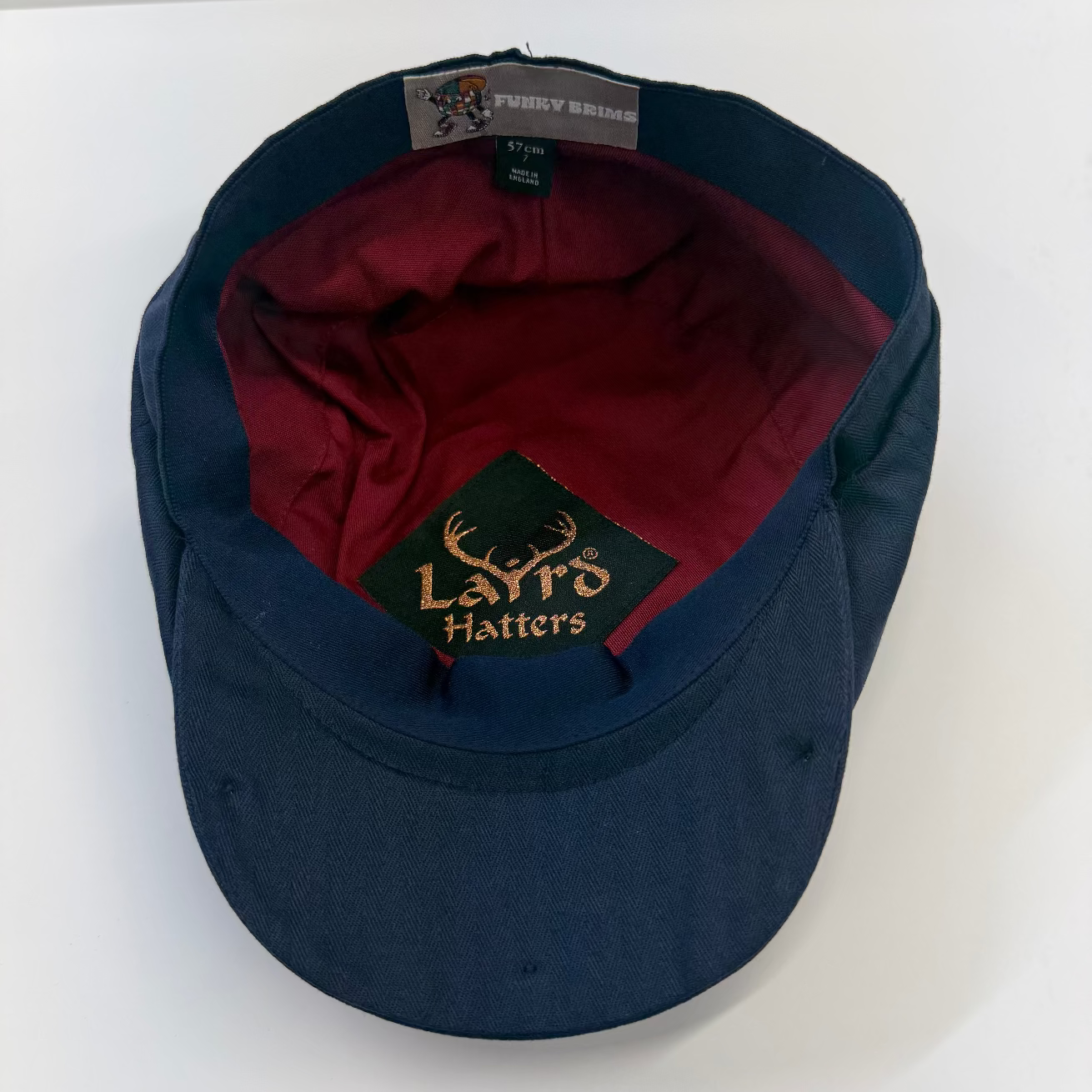 A navy blue snapback cap with a dark red interior lining, featuring a green embroidered patch on the front with gold text reading 'Laird Hatters' and a gray brand tag inside the crown.