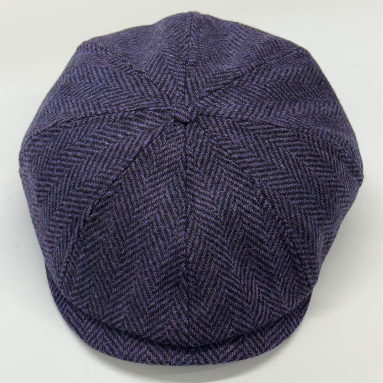 A dark purple herringbone pattern flat cap, also known as a newsboy cap, photographed from a top-down angle on a light gray background.