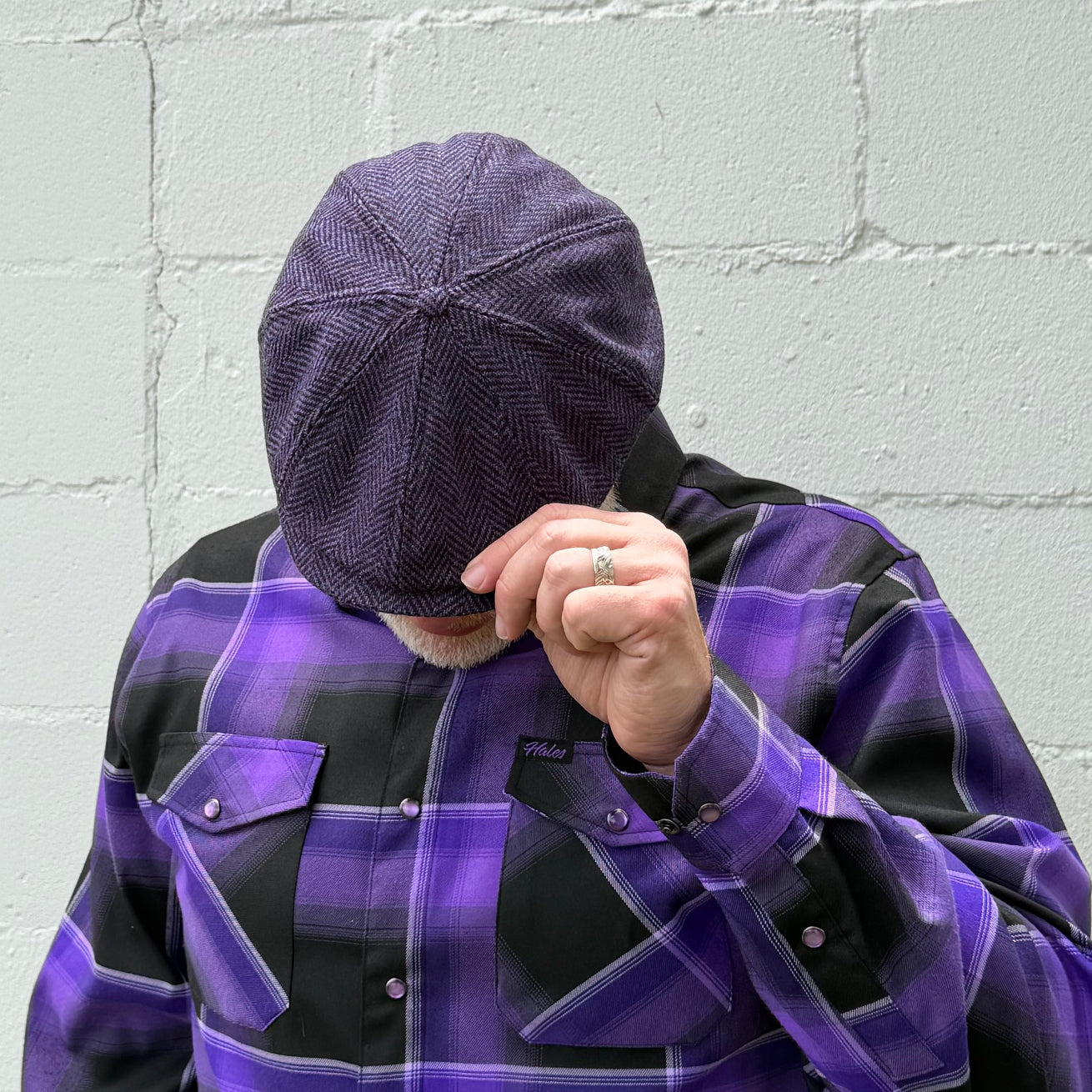 A person wearing a dark purple textured flat cap and a purple and black plaid button-up shirt, holding the brim of the cap with one hand in front of a light gray brick wall.
