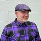A man wearing a purple and black plaid button-down shirt and a matching purple flat cap from FunkyBrims, standing against a light gray painted brick wall.