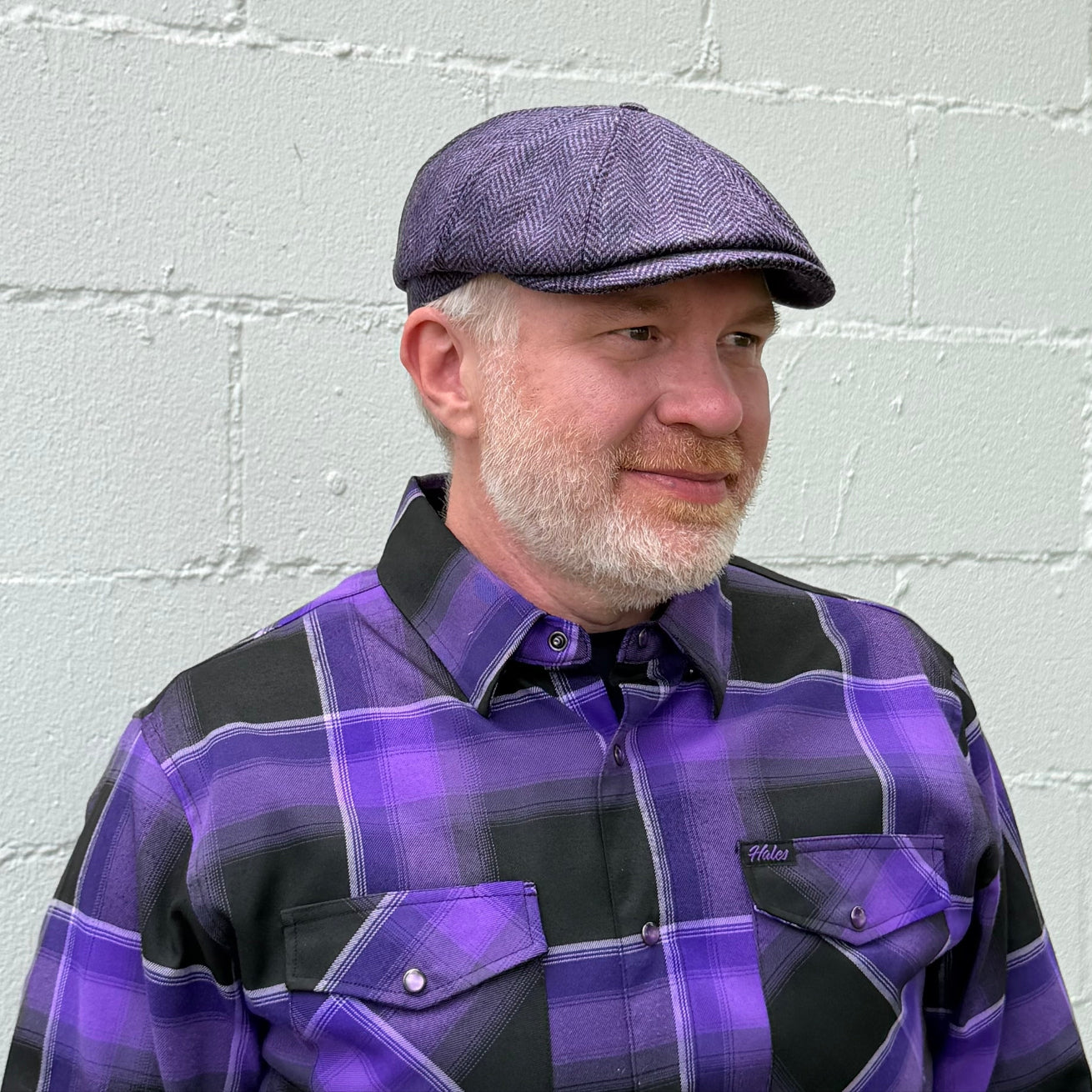 A man wearing a purple and black plaid button-down shirt and a matching purple flat cap from FunkyBrims, standing against a light gray painted brick wall.