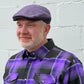 A man wearing a purple and black plaid button-down shirt and a matching purple flat cap, standing against a white painted brick wall.